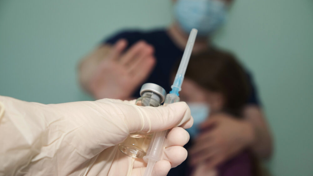 médico segura vacina e seringa em primeiro plano. Atrás, desfocado, pai protege filha, com a mão sinalizando que não quer a vacina