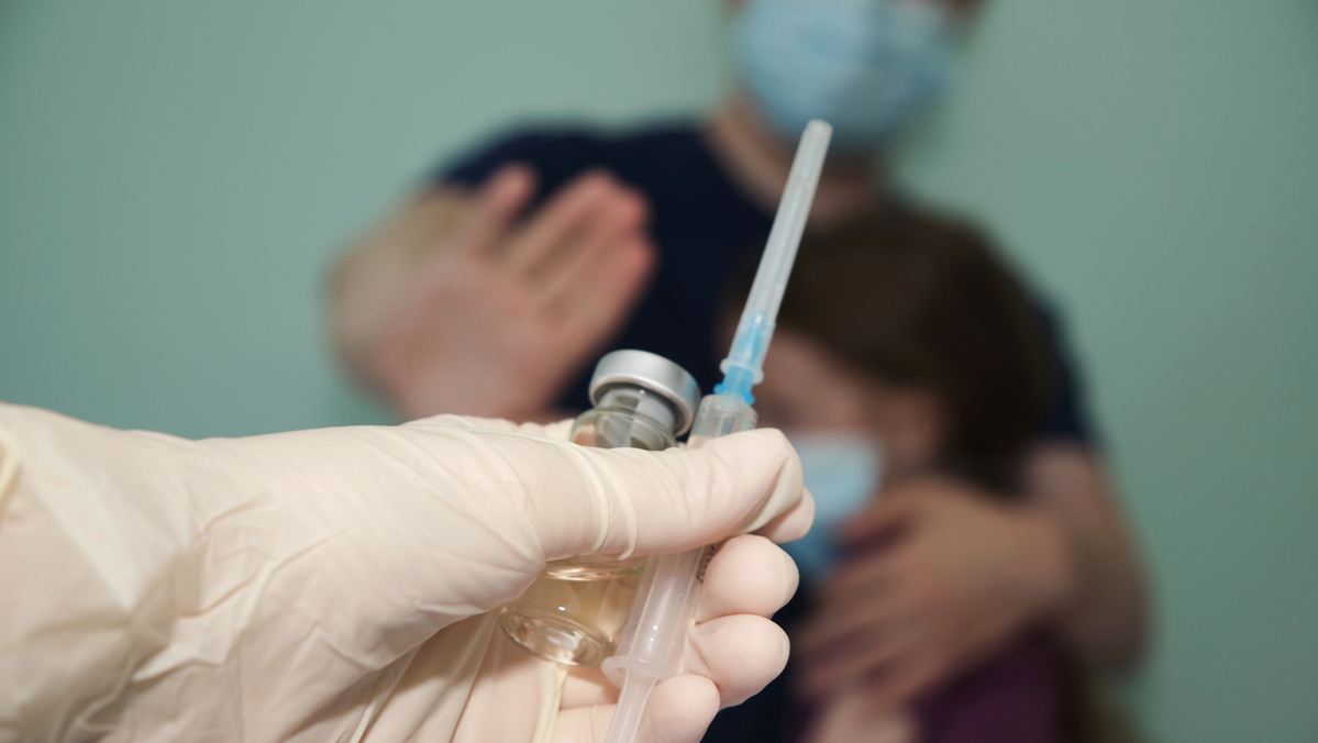 médico segura vacina e seringa em primeiro plano. Atrás, desfocado, pai protege filha, com a mão sinalizando que não quer a vacina