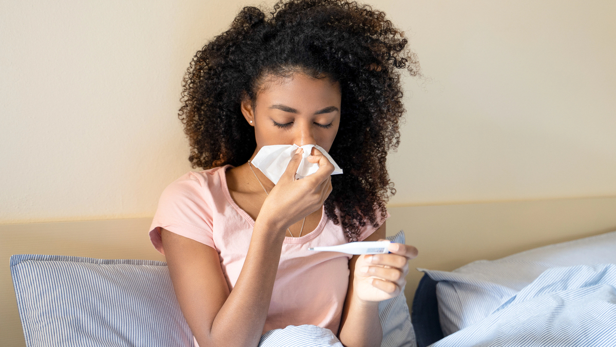 mulher de cama, com gripe, assoando o nariz e olhando o termômetro