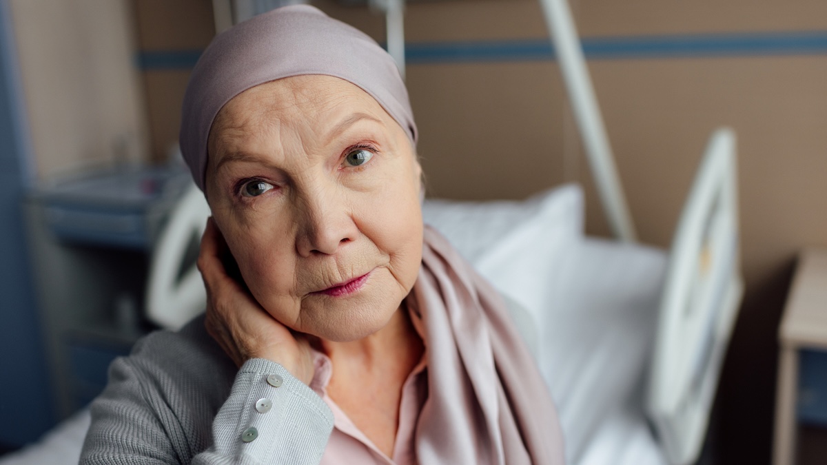 Older woman in a hospital bed wearing a pale pink headscarf, resting her hand at her neck and gazing to the side.