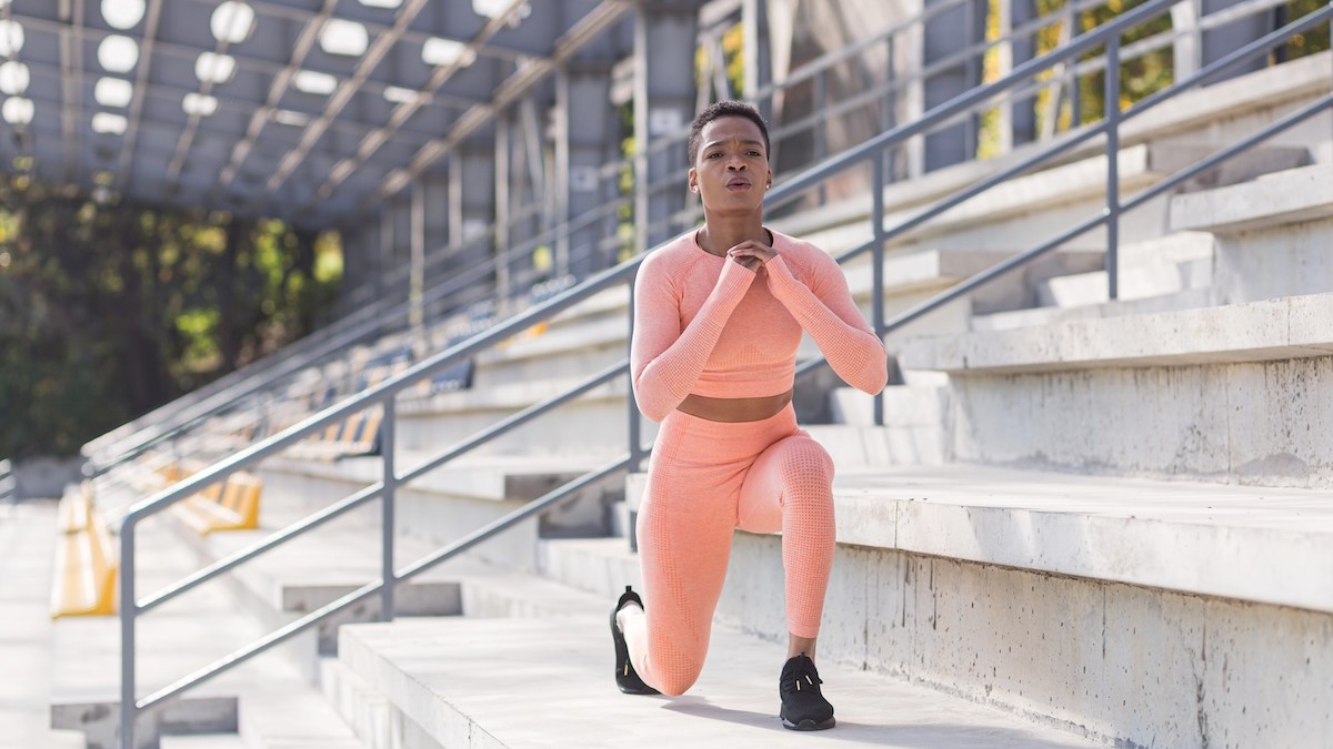 Athlete in peach workout outfit performing a lunge on stadium steps outdoors, one knee on a step and hands near chest.