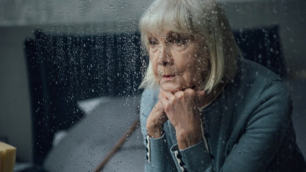 Older woman on a corded phone, resting her chin on her hands, seen through a rain-speckled window.