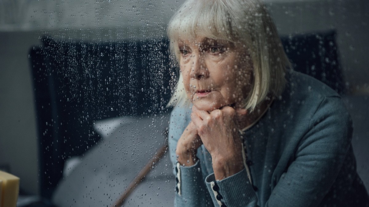 Older woman on a corded phone, resting her chin on her hands, seen through a rain-speckled window.