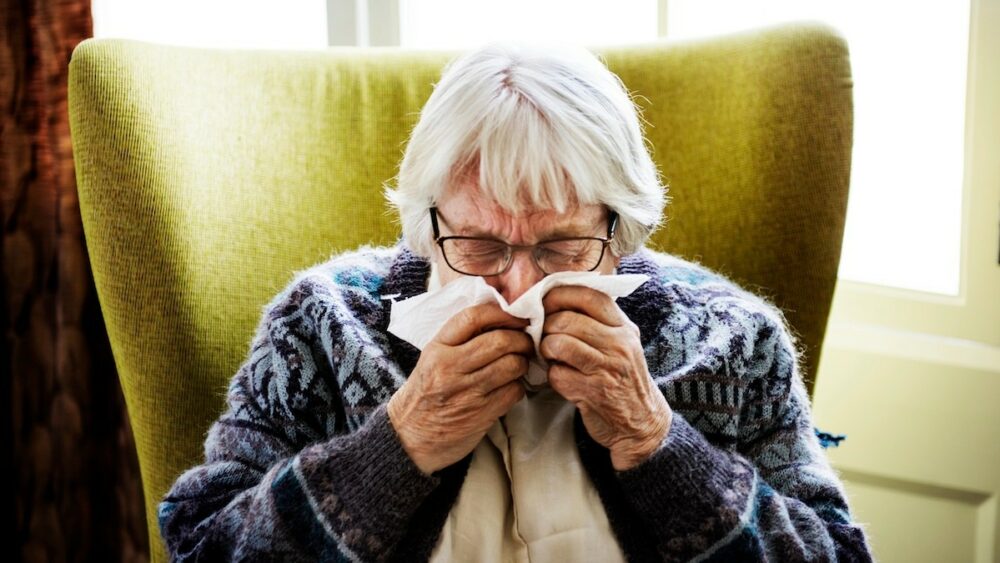 Elderly woman with short white hair and glasses blowing her nose into a tissue while seated in a green armchair.