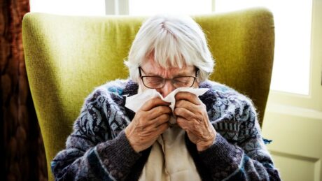 Elderly woman with short white hair and glasses blowing her nose into a tissue while seated in a green armchair.
