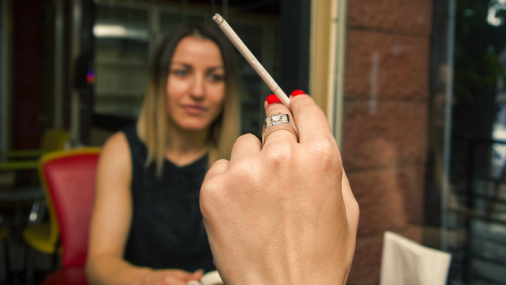 mão de mulher segurando cigarro diante de amiga fumante passiva