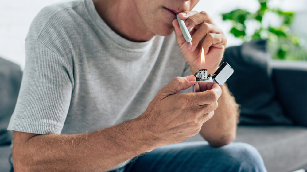 homem sentado em sofá da sala acende cigarro de maconha (cannabis)